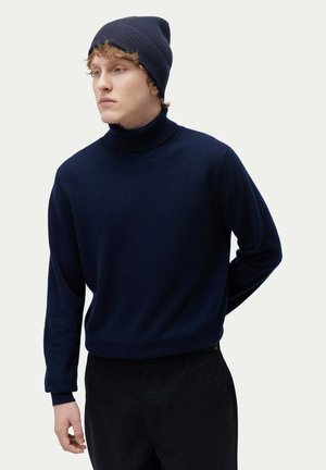 Navy turtleneck sweater made from soft material, fitted design with long sleeves, paired with a matching ribbed beanie and dark pants.