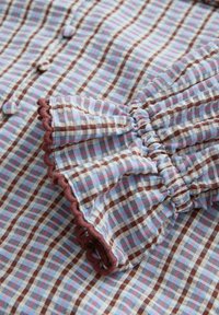 Prim-plan cu țesătură în carouri albastre, maro și roz, cu mansetă adunată și volanată, marginită cu tiv maro și nasturi acoperiți cu țesătură.