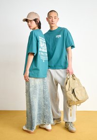 Teal oversized t-shirts with graphic prints, lightweight gray sweatpants, snake-patterned skirts, beige cap, and a tan backpack.