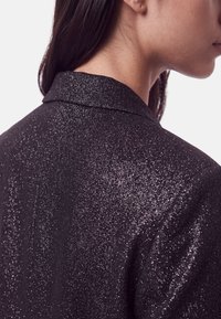 Femme portant une veste texturée sombre et étincelante vue de dos et de profil, avec de longs cheveux foncés et un teint clair.