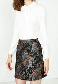 Vit långärmad blus med en rosett vid halsen, parat med en svart blommönstrad knä-lång kjol.