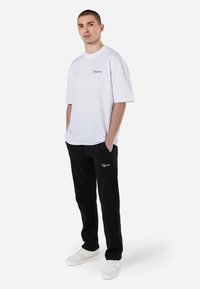 Balta per didelė t-shirt su apvaliu iškirpimu ir pilku logotipu, derinama su juodais šonkauliniais džinsais, turinčiais šonines kišenes ir siuvinėjimu, nešiojama su baltais sportbačiais.