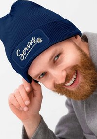 Navy blue knitted beanie with a textured surface, featuring a rectangular patch with the word "Servus" and a flower design.