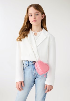 Młoda dziewczyna z długimi włosami, ubrana w biały krótki blezer i niebieskie dżinsy, niesie małą różową torebkę w kształcie serca na łańcuszku.