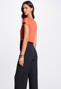 Boven: kortmouw, gestructureerde, oranje blouset. Onder: hoge taille, losvallende, zwarte broek. Model draagt gouden armbanden.