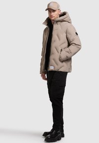 Giacca a vento in beige chiaro con design a trapuntato, cappuccio e dettagli neri. Abbinata a pantaloni cargo neri e stivali neri pesanti.