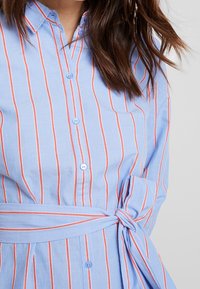 Jasnoniebieska sukienka na guziki w czerwone pionowe paski, z wiązanym w pasie fasonem, kołnierzykiem i gładką teksturą. Zawiera jasnoniebieskie guziki.