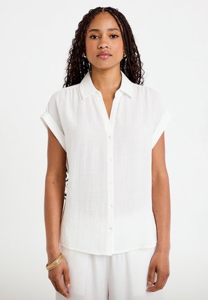 Femme aux longs cheveux tressés portant un chemisier blanc à manches courtes boutonné et un pantalon blanc sur un fond uni.