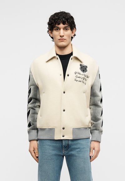 Άνδρας που φοράει μια μπεζ και γκρι varsity μπουφάν με μαύρα γραφικά κείμενα και λογότυπα, συνδυασμένο με μπλε τζιν, στέκεται μπροστά σε απλό φόντο.