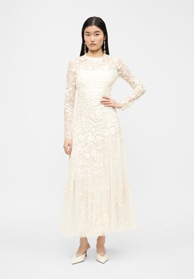 Γυναίκα που στέκεται με το ένα χέρι στη μέση, φορώντας ένα μακρύ, διάφανο λευκό φόρεμα με λουλουδένια δαντέλα και λευκές μυτερές γόβες που ταιριάζουν.