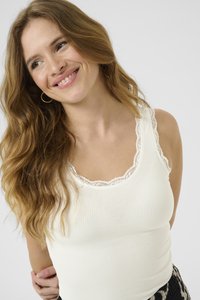 Top sin mangas blanco con ribetes y un escote adornado con encaje; combinado con pantalones oscuros de patrón; la modelo tiene el cabello castaño ondulado.