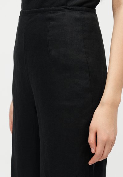 Μαύρο παντελόνι από λινό με λεία υφή και σχέδιο με φαρδιά μπατζάκια, που αναδεικνύει τη ψηλή μέση και λεπτομέρειες στη ραφή στο πλάι, με το χέρι να ακουμπά στο ισχίο.