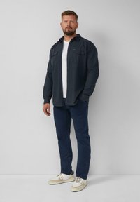 Camicia button-up blu scuro sopra una t-shirt bianca, abbinata a pantaloni navy e sneakers bianche con accenti beige. Tessuto liscio con una vestibilità classica.