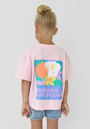 Dziecko z blond włosami spiętymi w kok, ubrane w różowy T-shirt z kolorowymi kwiatami i napisem "ROŚNIĘ DZIEŃ PO DNIU" na plecach oraz w jeansowe spodenki.