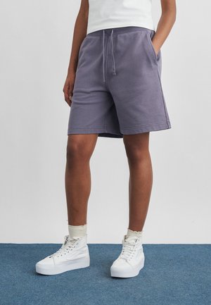 Persona que lleva puestos unos pantalones cortos grises con cordón, calcetines blancos y zapatillas blancas de caña alta, de pie sobre una alfombra azul.