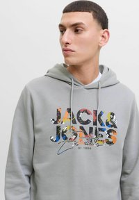 Sudadera gris con un colorido estampado gráfico de "JACK & JONES Essentials" en el pecho. Capucha con cordones y textura suave.