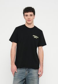 T-shirt de algodão preta com mangas curtas e gola redonda. Texto gráfico amarelo "FRUITS OF LOVE" no lado esquerdo do peito. Textura macia.