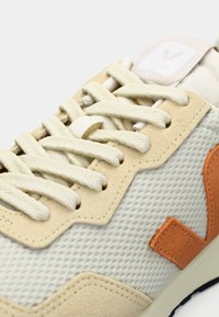 Beige mesh-sneaker med semsket skinn-detaljer, kontrasterende oransje detaljer og flate lisser. Inkluderer en polstret krage og logo på tungen.