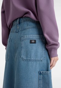 Pantalones cortos de mezclilla de color azul claro con dos bolsillos traseros, que presentan una etiqueta negra de Vans. El conjunto se complementa con una camiseta de manga larga morada.