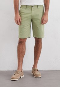 Light green cotton shorts with a relaxed fit, featuring a button closure and pockets. Paired with beige suede shoes.