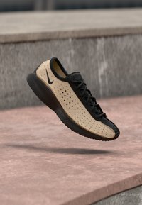 Μαύρο και μπεζ αθλητικό παπούτσι Nike με διάτρητες πλευρές και μαύρη σόλα, εμφανιζόμενο στον αέρα πάνω από μια ρόζ τσιμεντένια επιφάνε ια.