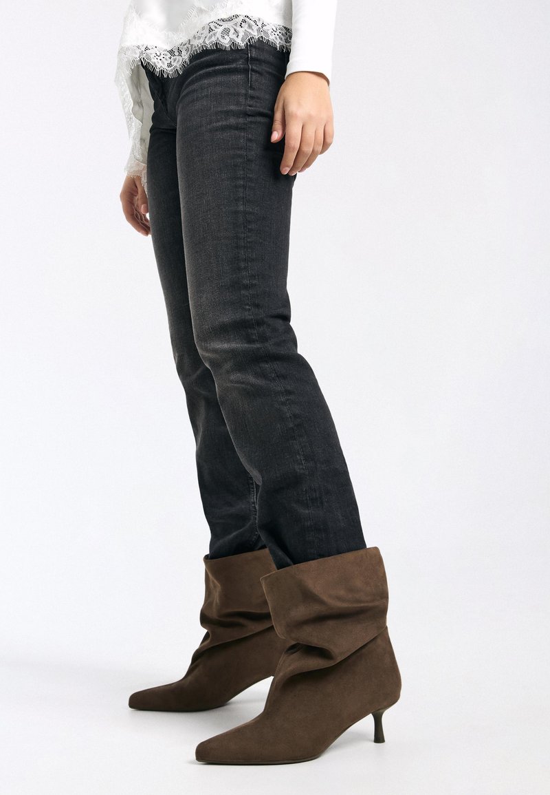 Stivaletti alla caviglia in suede marrone con design morbido e punte appuntite, abbinati a jeans in denim scuro e una maglia bianca con dettagli in pizzo.