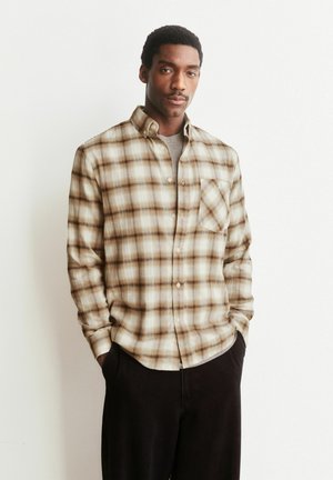 Mann trägt beige und braunes kariertes Hemd mit Knöpfen, graues Unterhemd und schwarze Hose, steht mit Händen in den Taschen vor weißem Hintergrund.