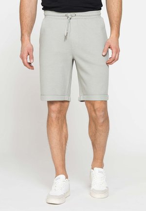 Uomo con pantaloncini grigio chiaro con coulisse e sneakers bianche in piedi davanti a uno sfondo semplice.
