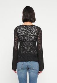 Μαύρο πουλόβερ από δαντέλα με μακριά μανίκια, που διαθέτει περίπλοκο διαμάντι μοτίβο, άνετη εφαρμογή και ριμπ μανσέτες. Φορεμένο πάνω από μπλε τζιν.