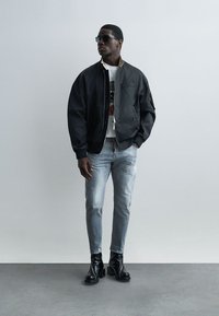Homme portant une veste bomber noire, un T-shirt blanc à motif, un jean bleu clair déchiré, des bottes à talons noires et des lunettes de soleil, debout contre un mur gris uni.
