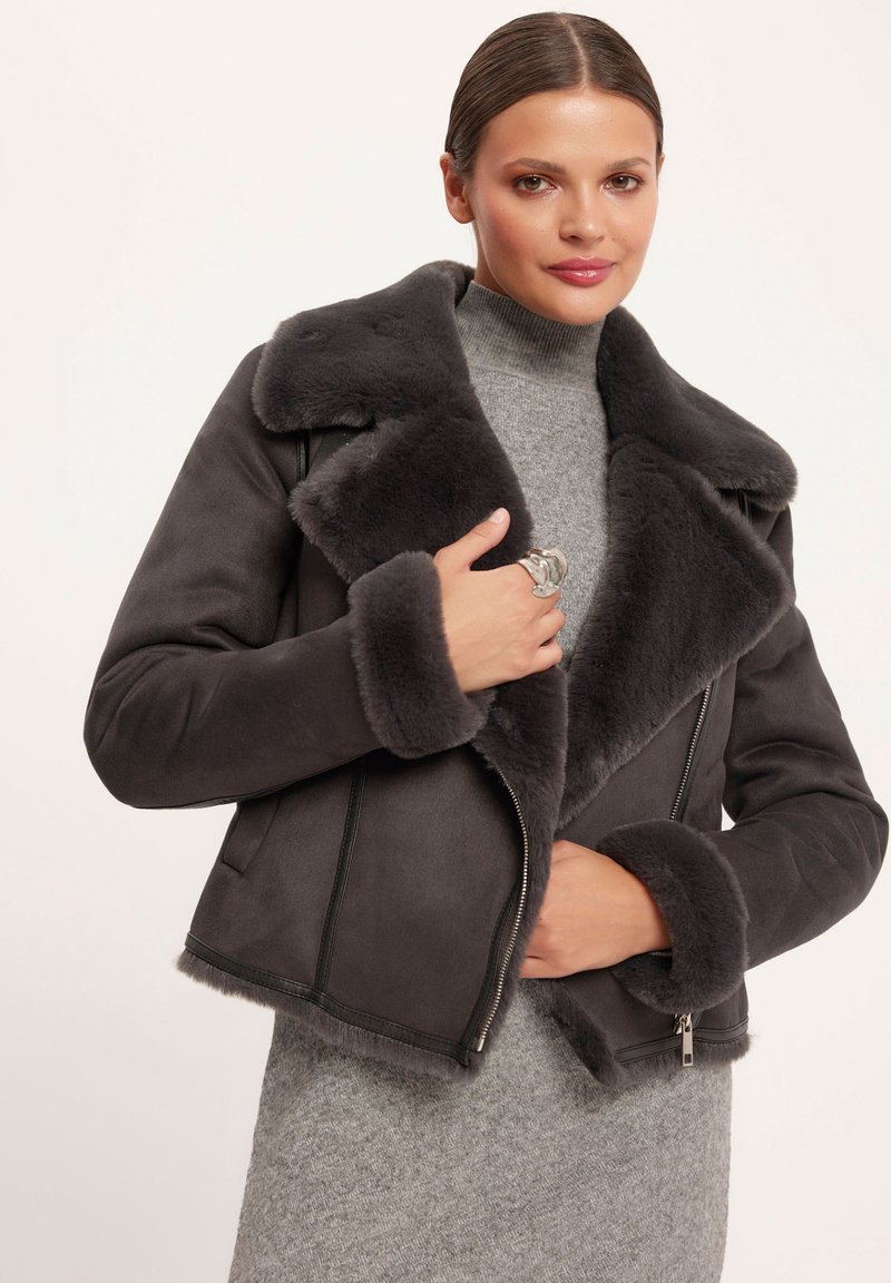 Szary sztuczny żakiet z kożuchem z szerokim kołnierzem, zamknięciem na zamek i puszystym wykończeniem na mankietach. Noszony na jasnoszarym golfie.