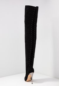 Bottes noires cuissardes en daim doux. Dotées d'un détail lacé sur le côté et d'un talon haut métallique. Texture lisse sans motifs visibles.