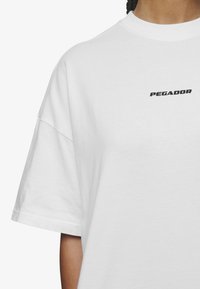 Bijela pamučna majica s crnim logotipom "PEGADOR" na prsima. Majica ima širok kroj, spuštena ramena i kratke rukave.