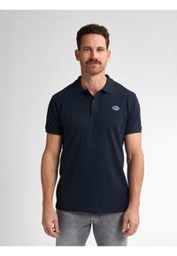 Tumman sininen polo-paita puuvillakangasta, jossa on kaulus, lyhyet hihat ja pieni, brodeerattu logo rinnassa. Yhdistetty harmaisiin farkkuihin.