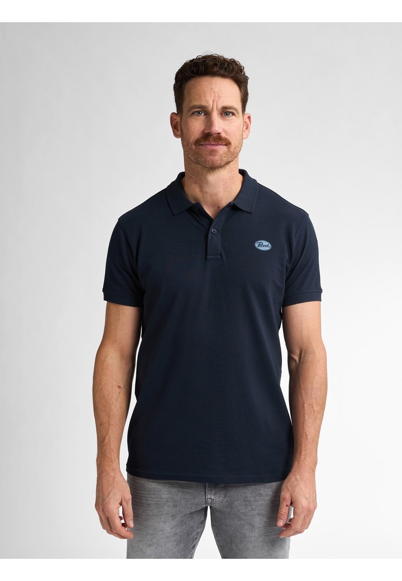 Tumman sininen polo-paita puuvillakangasta, jossa on kaulus, lyhyet hihat ja pieni, brodeerattu logo rinnassa. Yhdistetty harmaisiin farkkuihin.