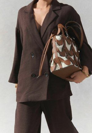 Personne portant un costume marron texturé tenant un sac à main beige et marron à motifs avec des anses en cuir sur fond clair.