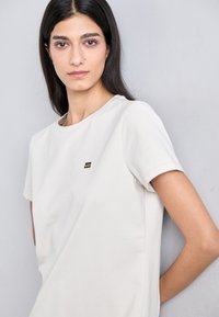 Jasnobeżowa koszulka z krótkim rękawem, uszyta z gładkiego materiału, z małym czarnym logo na piersi. Prosty, klasyczny design z okrągłym dekoltem.