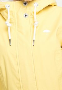 Imperméable jaune avec cordons de serrage blancs, perles en bois, boutons-pression en métal et petit logo de parapluie blanc sur la poitrine.
