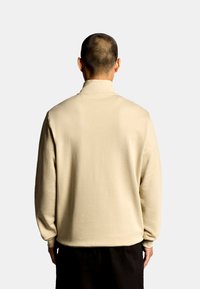 Beiger Pullover-Sweatshirt mit hohem Kragen, langen Ärmeln und glatter Textur, von hinten vor hellem Hintergrund gesehen.