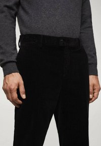 Pantaloni in corduroy neri con una texture a coste, dotati di chiusura a bottone e design della vita. È visibile la maglietta a maniche lunghe grigia del modello.