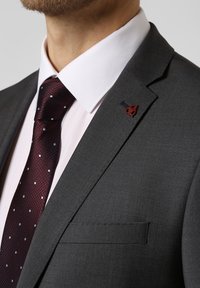 Szary żakiet w stylu garsonki z klapami, biała koszula elegancka i bordowa krawat w kropki. Zauważalny detal z czerwono-czarną przypinką na klapie.