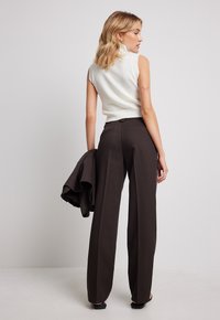 Un pull à col roulé sans manches blanc associé à un pantalon marron taille haute et à pattes d'éléphant. Le mannequin tient une veste sombre. Textures de tissu lisses.