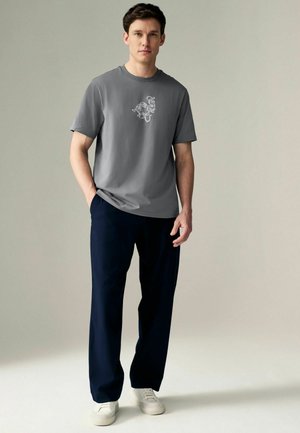 T-shirt grise à manches courtes avec imprimé graphique sur la poitrine, associé à un pantalon large bleu marine et des baskets blanches sur un fond neutre.
