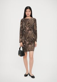 Μακρυμάνικο μπεζ φόρεμα με καφέ μπορντούρα, συνδυασμένο με μαύρη τσάντα και μαύρα φλατ παπούτσια. Λείο ύφασμα με εφαρμοστό σχήμα.