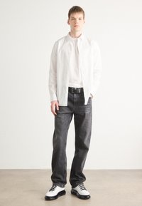 White button-up shirt over a light top, paired with dark jeans. Black belt and white shoes with black accents. Simple design, casual fit.