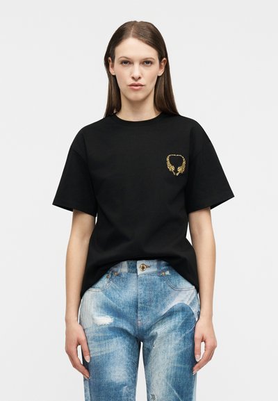 Femme aux cheveux bruns raides portant un t-shirt noir avec un petit motif de tigre doré et un jean bleu clair délavé, debout, droite.