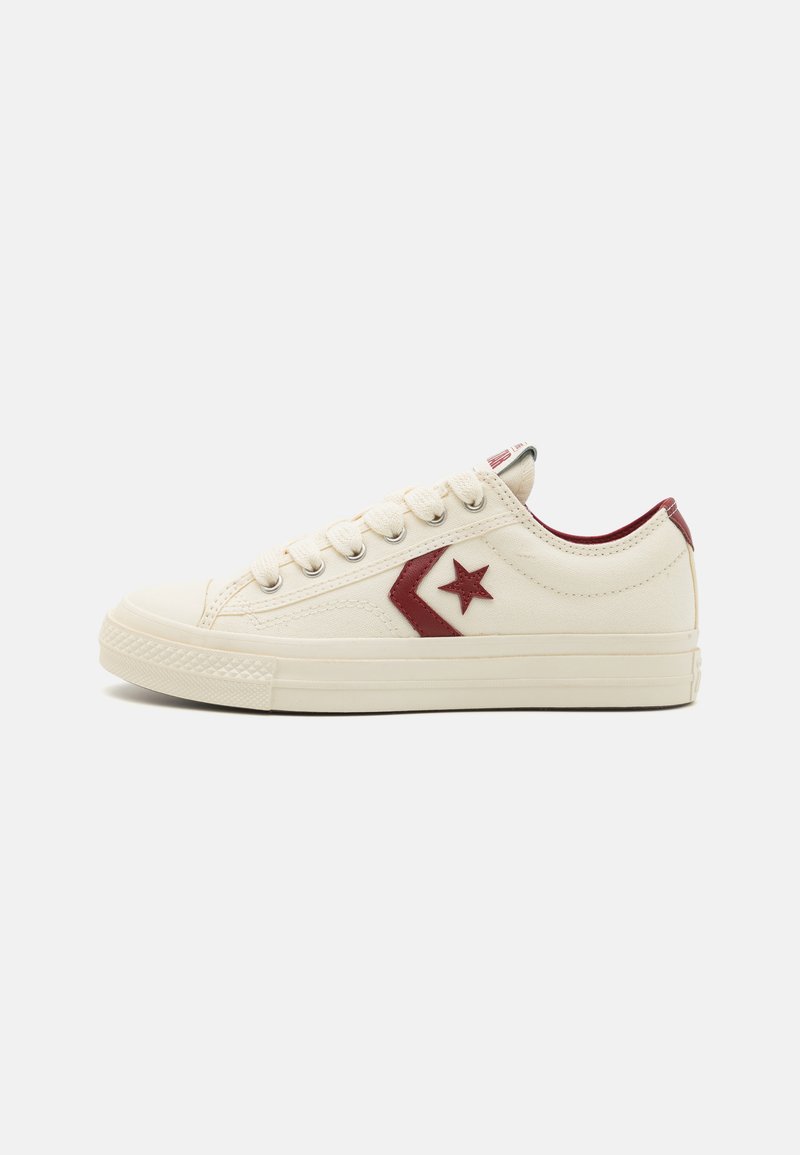 Canvas sneakers in cream, featuring a red star logo and accent. Round toe, lace-up front, and textured sole. Side view.