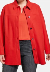 Chaqueta de tela suave roja con cuello, botones frontales y bolsillos laterales, superpuesta sobre una camiseta naranja y jeans oscuros.