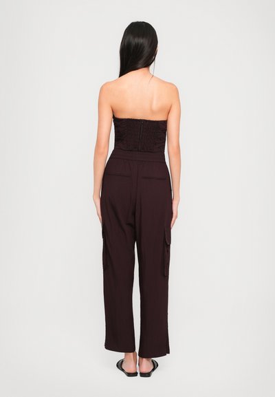 Top bez naramenica u boji burgundca s smock teksturom i skrivenim patentnim zatvaračem, uparen s hlačama širokih nogavica koje imaju bočne džepove i gumbe kao ukras.