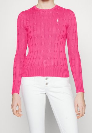 Femme portant un pull à torsades rose vif et un pantalon blanc taille haute avec trois boutons visibles.